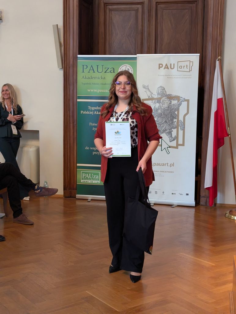 Lic. Zuzanna Śliwa z dyplomem na tle plakatu promującego wydarzenie